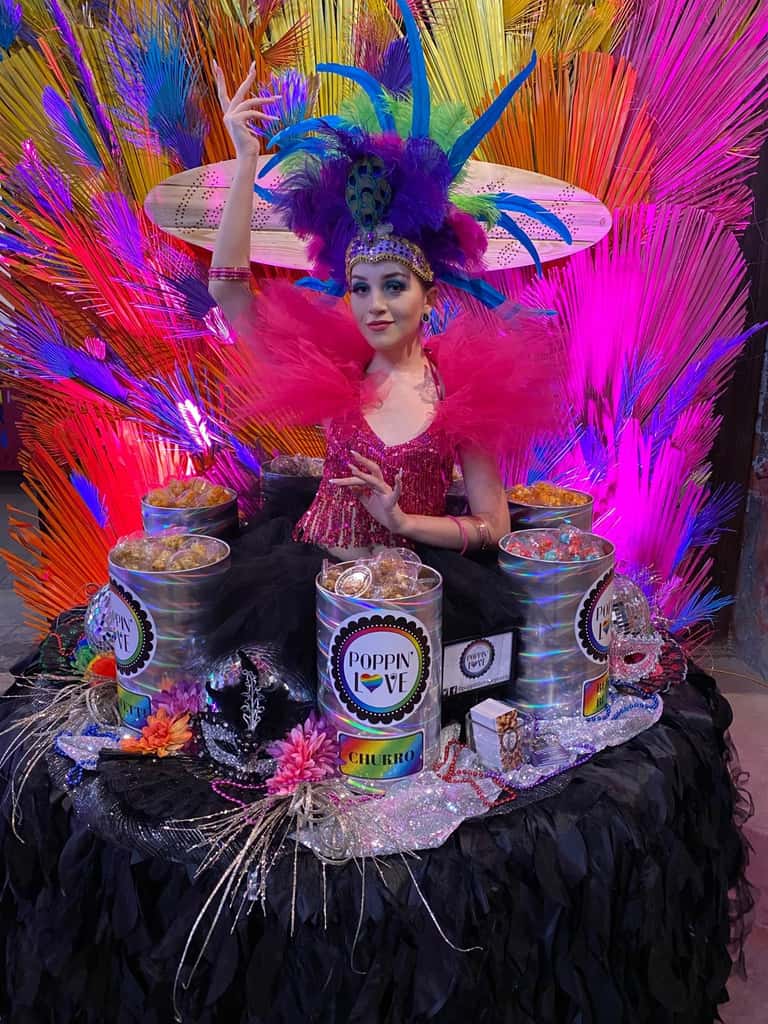 A Carnivale performer in vivid rainbow plumage beside a festive display