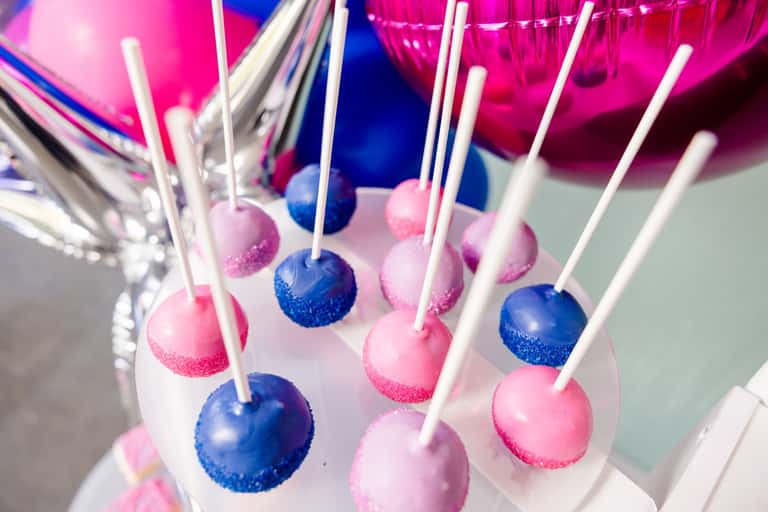 Hand-dipped cake pops in pink, violet, and blue, rimmed with sanding sugar on a tiered stand
