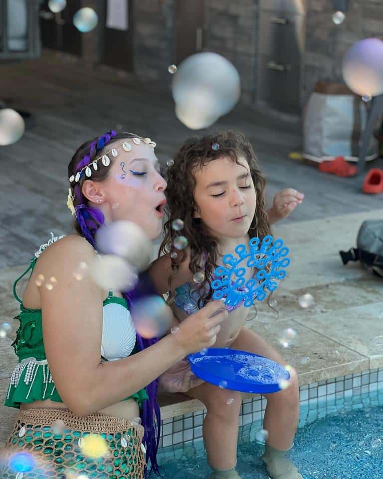 A mermaid performer blows bubbles with a child at a poolside party