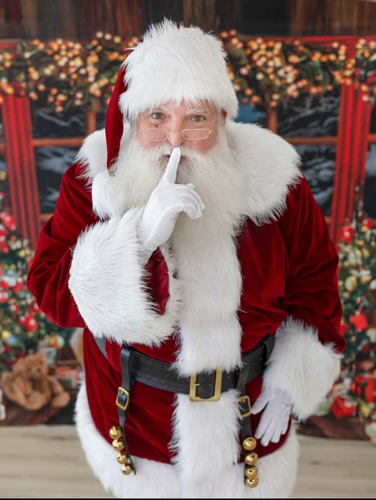 Santa holds a white-gloved finger to his lips, hushing the room before the surprise