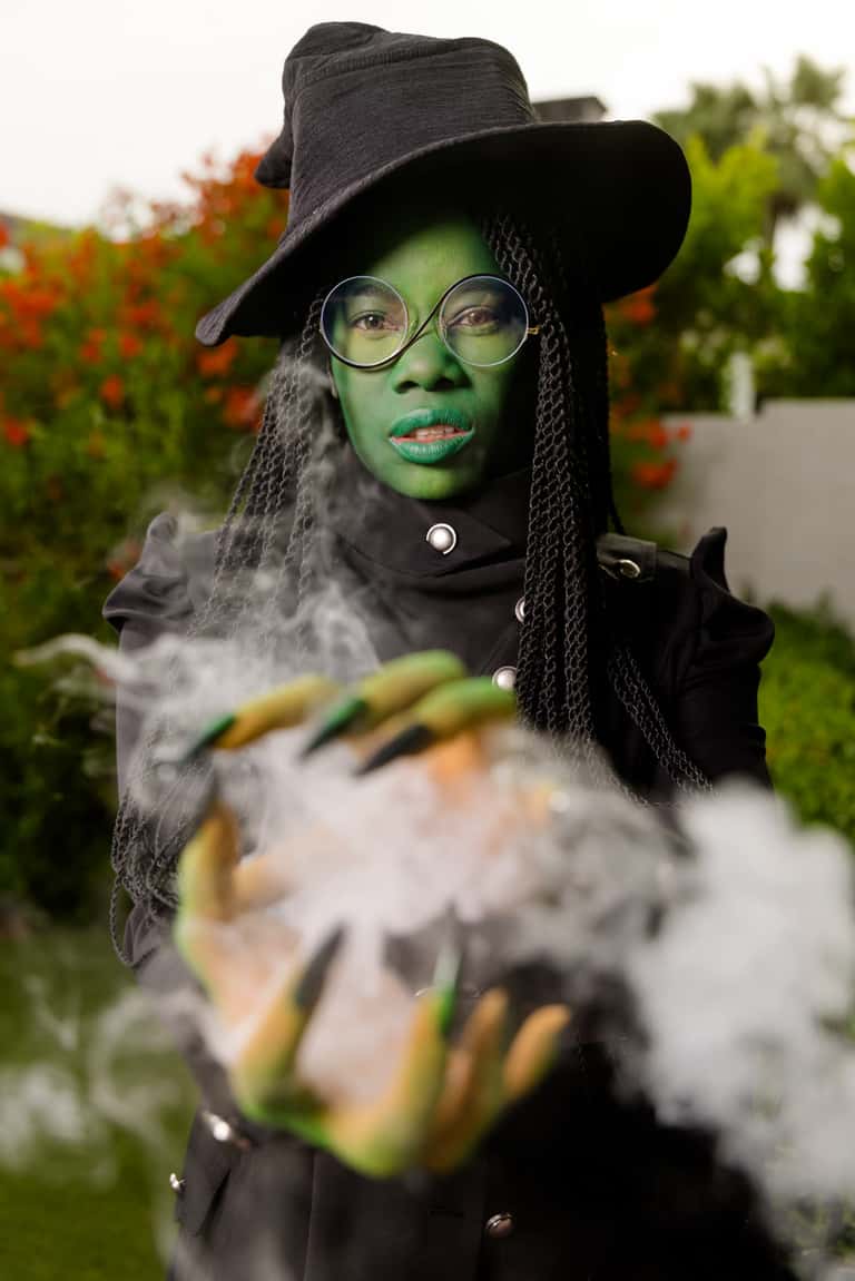A theatrical green-skinned witch character conjures stage fog
