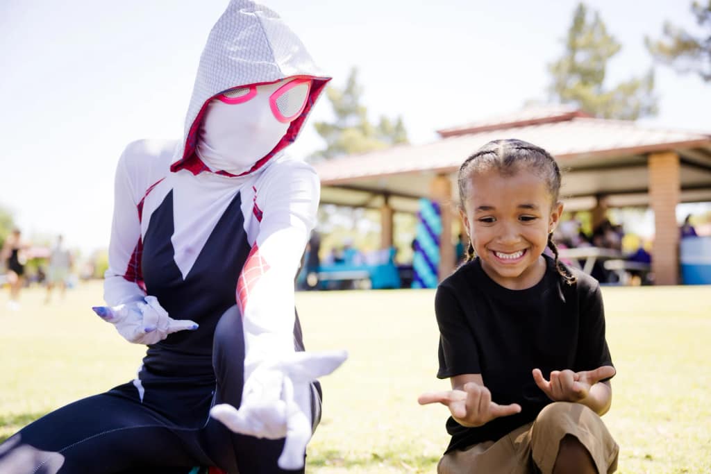 A Spider-Gwen character shares a playful moment with a young guest on a lawn